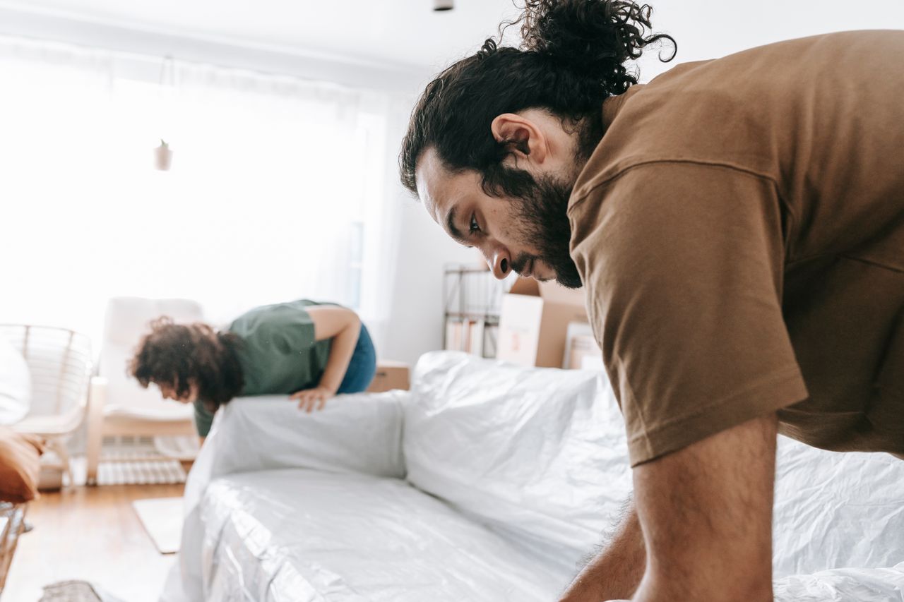 A couple wrapping furniture in a bright apartment, getting ready for a move.